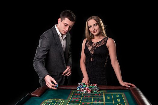 Elegant couple at the casino betting on the roulette, on a black background. A man in a suit with a beautiful young woman in a black dress