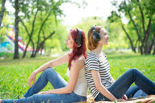 Two young women listen to music in the park. Sitting back to back