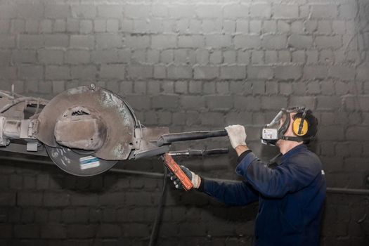 A man in work clothes and a respirator, controls heavy equipment with grinding stone to clean reinforced concrete structures in a workshop of an industrial plant.