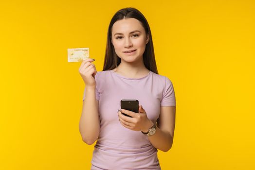 Portrait of a happy young blonde girl showing plastic credit card while holding mobile phone isolated over yellow background - Image.