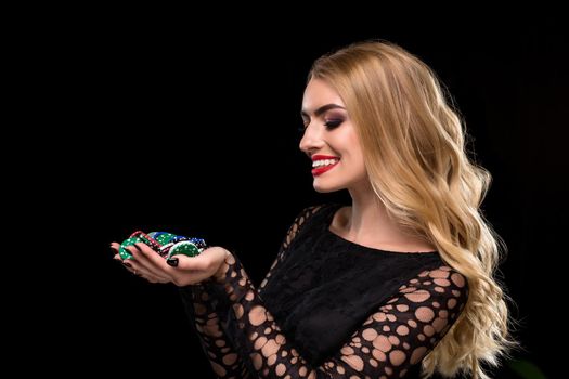 Elegant blonde in a black dress, casino player holding a handful of chips on black background. Poker. Casino. Roulette Blackjack Spin. Caucasian young woman looking at the chips emotions