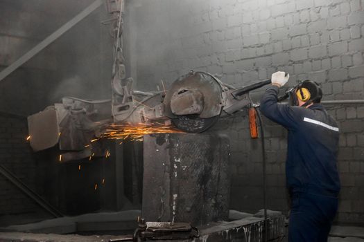 A man in a working jumpsuit and respirator, cleans cast iron tubing with heavy equipment with a grinding stone in the workshop of an industrial plant.