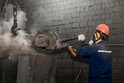 A worker in a protective helmet, respirator and overalls grinds with heavy equipment cast iron concrete tubing in the workshop of an industrial plant.
