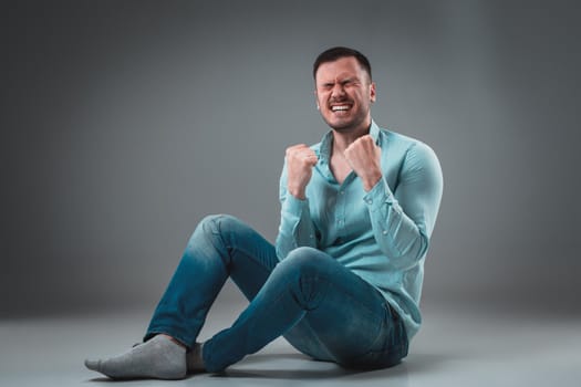 Appealing casual young man sitting on the floor, looking to the camera smiling. Studio shot.