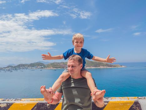 Front boy son laugh sitting on fathers shoulders. No photoshop sun on skin. Sea, clouds, island background. Funny photo, happiness lifestyle, Father's Day, love parenthood, family holiday concept.