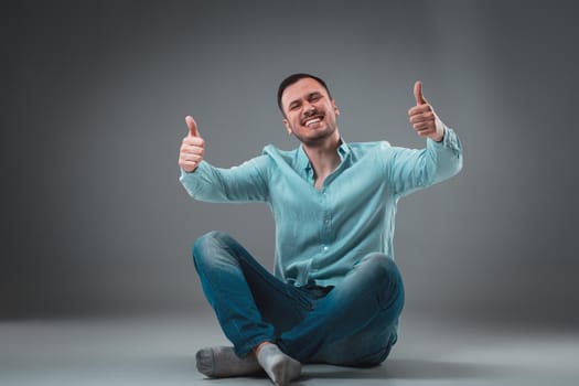 Handsome young man sitting on a floor with raised hands, isolated on gray background. Emotion concept