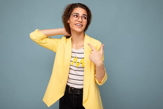 Photo of young positive happy adorable attractive brunette curly woman with sincere emotions wearing stylish yellow jacket and optical glasses isolated on blue background with copy space.