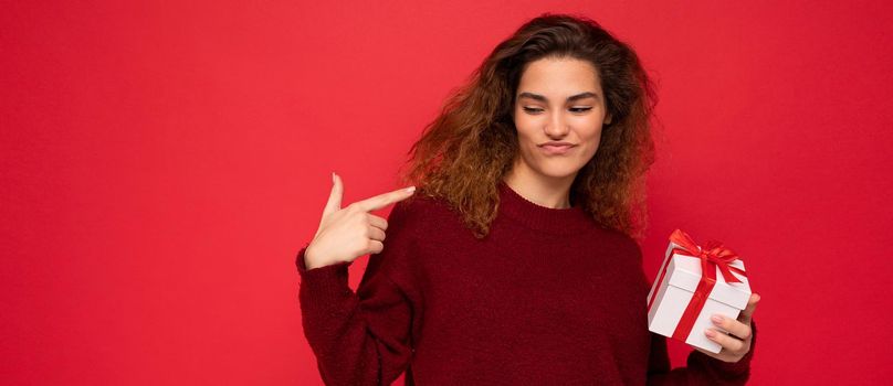Attractive positive self-confident emotional young brunette curly woman isolated over red background wall wearing red sweater holding gift box looking to the side.