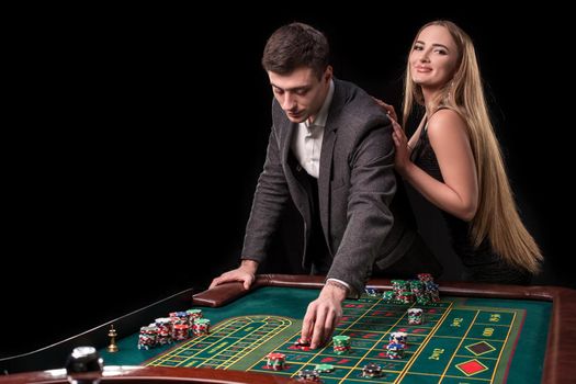 Elegant couple at the casino betting on the roulette, on a black background. A man in a suit with a beautiful young woman in a black dress