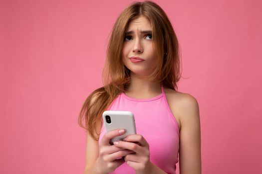Closeup photo of beautiful thoughtful young blonde woman wearing pink top poising isolated on pink background with empty space holding in hand and using mobile phone surfing on the internet online looking to the side and having doubts.