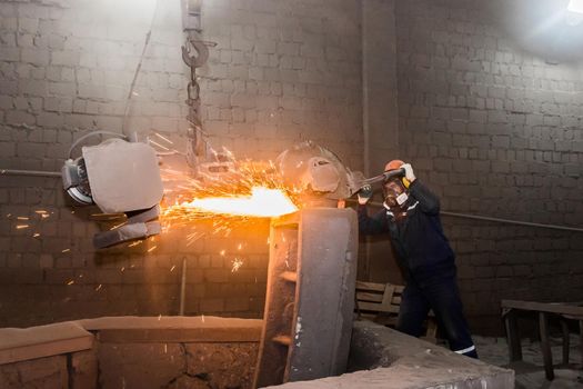 A male worker in a protective helmet, respirator, overalls manages heavy grinding equipment for cast iron concrete tubing with flying sparks in the workshop of an industrial plant.