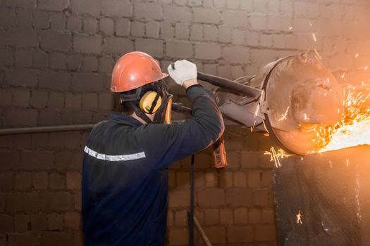 A male worker in a protective helmet, respirator, overalls manages heavy grinding equipment for cast iron concrete tubing with flying sparks in the workshop of an industrial plant.