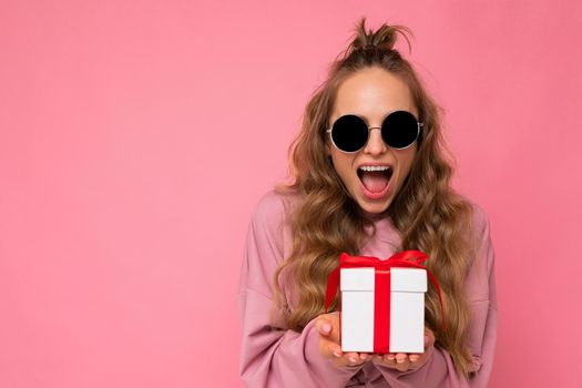 Photo shot of pretty positive surprised young blond woman isolated over colourful background wall wearing trendy outfit look holding gift box and looking at camera. Copy space