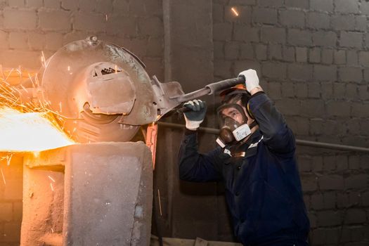 A male worker in a protective helmet, respirator, overalls manages heavy grinding equipment for cast iron concrete tubing with flying sparks in the workshop of an industrial plant.