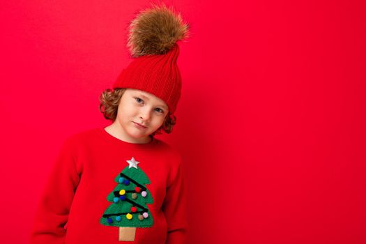 cool blond kid in warm hat and sweater with christmas tree on red background fooling around, christmas concept.