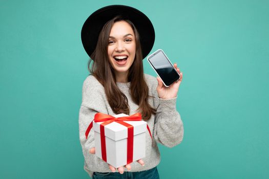 Photo of beautiful happy joyful young brunette woman isolated over blue background wall wearing stylish black hat and grey sweater holding gift box showing mobile phone screen display for mockup and looking at camera. Copy space, mockup