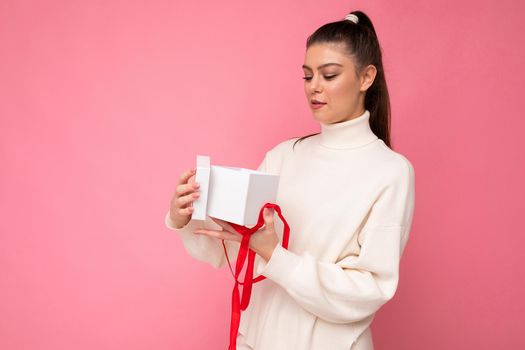 Attractive happy amazed young brunette woman isolated over pink background wall wearing white sweater holding gift box and unboxing present looking at camera.