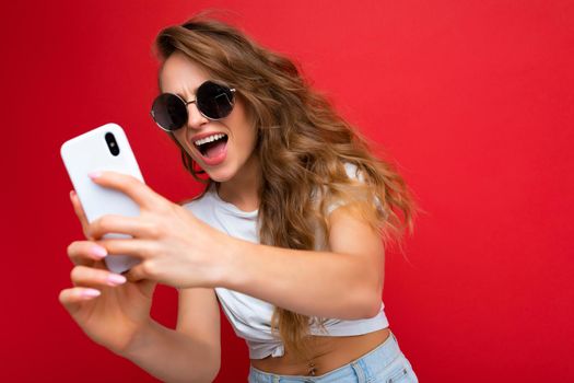 Attractive charming young smiling happy woman holding and using mobile phone taking selfie wearing stylish clothes isolated over wall background.