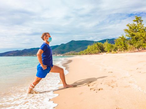 Coronavirus COVID 19. Young beautiful man doing sport performing workouts exercises and running near sea in the morning during quarantine. Active life in surgical sterilizing face mask protection.