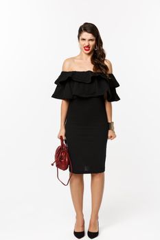 Beauty and fashion concept. Full length of angry woman in black party dress and high heels, express disdain and grimacing at camera, mad at person, white background.