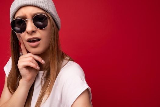 Closeup of young dissatisfied thoughtful emotional beautiful dark blonde woman with sincere emotions wearing casual white t-shirt, gray hat and sunglasses isolated on red background with copy space and thinking.