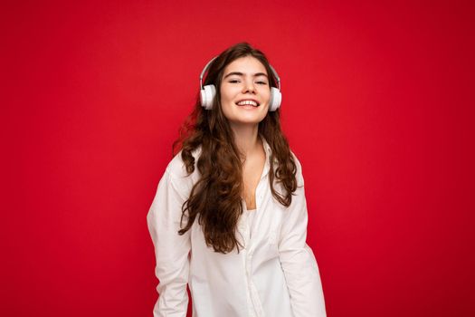 Attractive happy smiling young brunette female person wearing white shirt and optical glasses isolated over red background wearing white wireless bluetooth earphones listening to music and looking at camera.