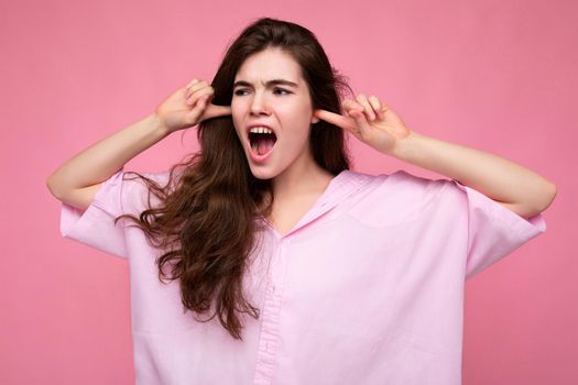 Portrait of young emotional winsome nice curly brunette woman with sincere emotions wearing casual pink shirt isolated over pink background with copy space and covering ears with fingers and shouting.
