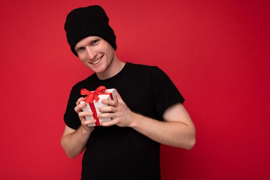 Shot of handsome positive smiling young man isolated over red background wall wearing black hat and black t-shirt holding white gift box with red ribbon and looking at camera.