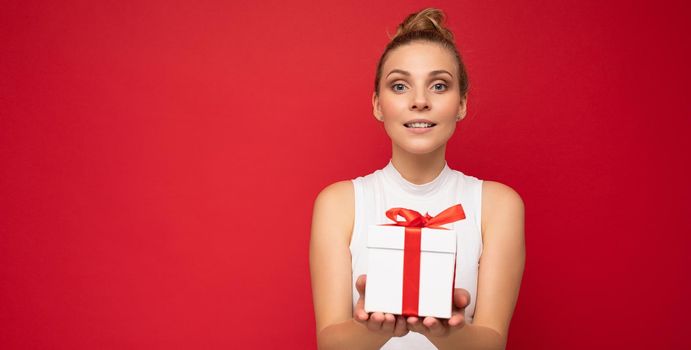 Beautiful happy young blonde woman isolated over colourful background wall wearing stylish casual clothes holding gift box and looking at camera. Empty space