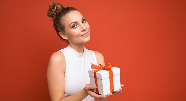 Pretty happy young blonde female person isolated over red background wall wearing white top holding gift box and looking at camera. Empty space