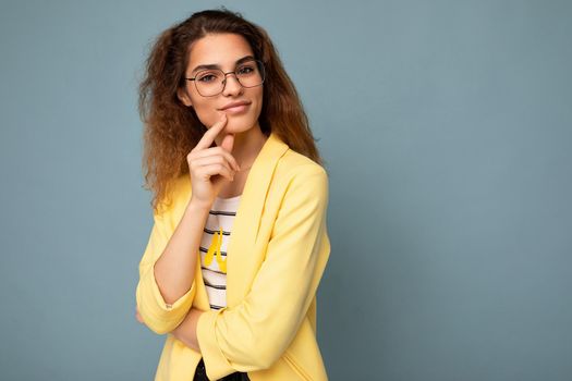 Photo of young positive thinking dreaming beautiful brunette curly woman with sincere emotions wearing casual yellow jacket and optical glasses isolated on blue background with copy space.