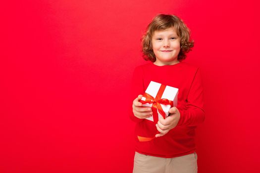 Photo shot of joyful smiling blonde curly boy isolated over red background wall wearing red sweater holding gift box and looking at camera. Copy space