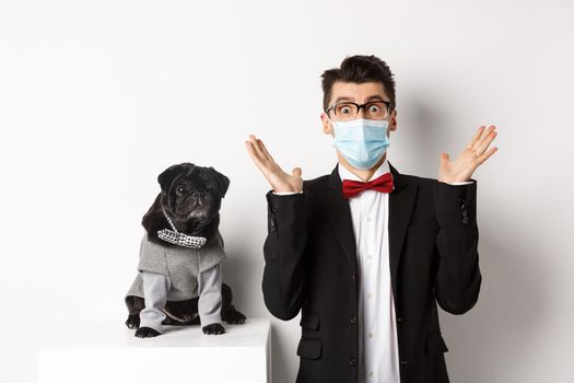 Coronavirus, pets and celebration concept. Amazed young man in face mask and suit staring at camera surprised, cute black dog sitting near owner in party outfit, white background.