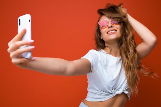 Attractive charming young smiling happy woman holding and using mobile phone taking selfie wearing stylish clothes isolated over wall background.