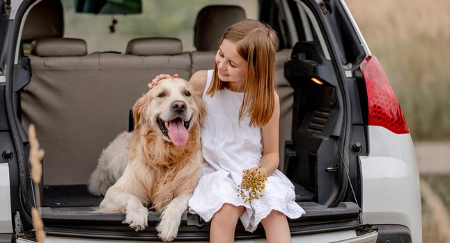 Preteen girl with golden retriever dog sitting in car trunk together. Pretty child kid hugging doggy pet in vehicle and looking at animal