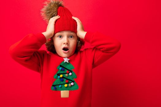 cheerful funny boy on a red background in a warm hat and sweater with a Christmas tree.