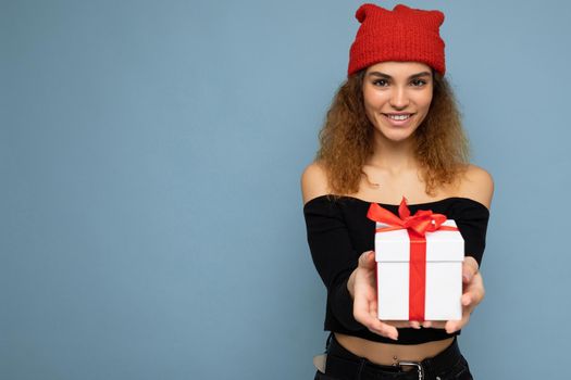 Beautiful happy young brunette woman isolated over colourful background wall wearing stylish casual clothes holding gift box and looking at camera. Empty space