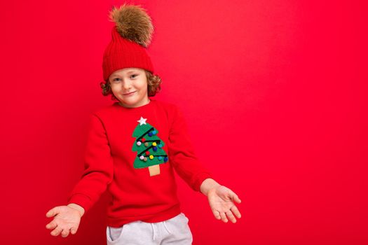 cool teenager in a red Christmas sweater fooling around against the background of a red wall, a warm hat and a sweater with a Christmas tree.