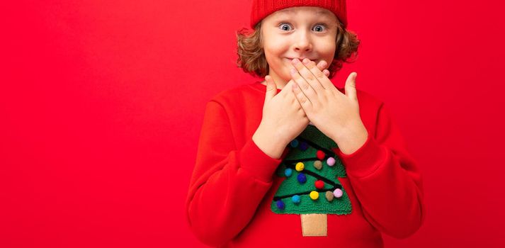 cool teenager in a red Christmas sweater fooling around against the background of a red wall, a warm hat and a sweater with a Christmas tree.