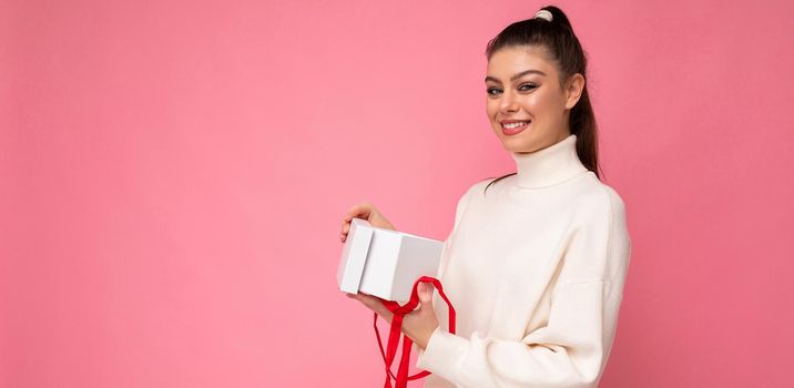 Attractive happy positive smiling young brunette woman isolated over pink background wall wearing white sweater holding gift box and unboxing present looking at camera. Copy space, mockup