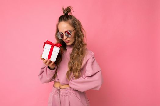 Attractive happy adult blonde curly woman isolated over pink background wall wearing pink sport clothes and sunglasses holding gift box looking at present. Copy space, mockup
