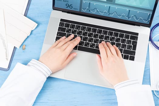 Top view of doctor hands typing at laptop computer. Therapist in white coat sitting at blue wooden desk. Examination and consultation in clinic. Medical application and online healthcare services.