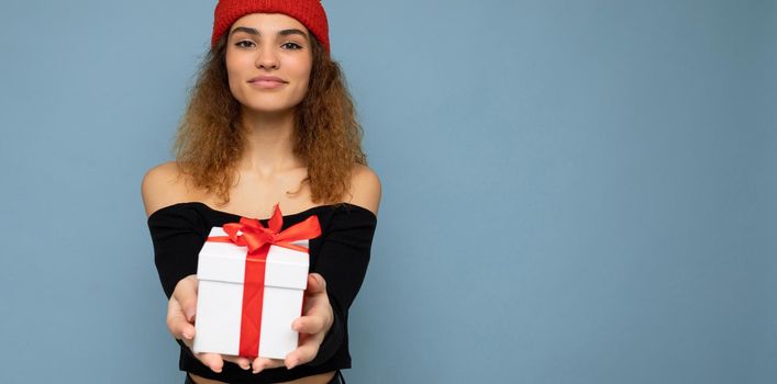 Shot of beautiful positive smiling young dark blonde curly woman isolated over blue background wall wearing black crop top and red hat holding gift box looking at camera. Free space