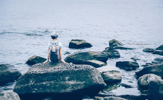 Traveler young woman with backpack sitting on big stone in the sea in summer, rear view. Image with instagram color effect