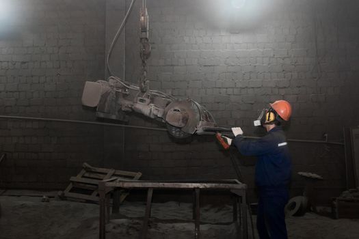 A working man in overalls, a protective helmet and a respirator controls heavy equipment for cleaning cast iron concrete tubing in the workshop of an industrial plant.