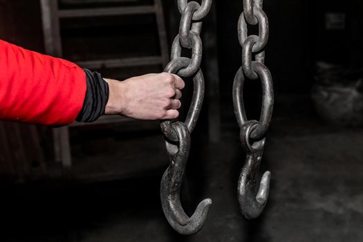 The hand of a male worker holds an iron chain with a hook lifting mechanism of a crane in an industrial plant.