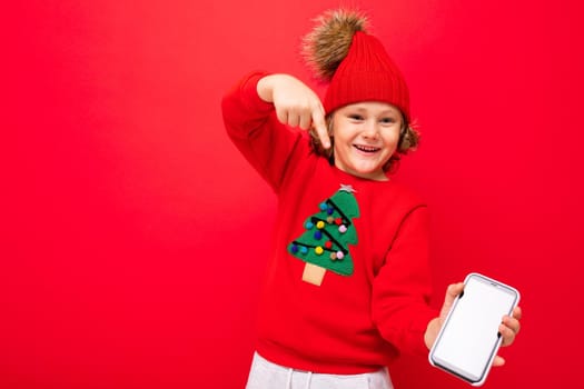 a young man with a smartphone in his hands against the background of a red wall, in a knitted hat and a sweater with a Christmas tree