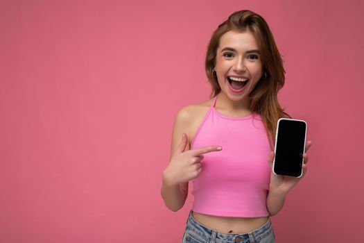 Photo of beautiful smiling young woman good looking wearing casual stylish outfit standing isolated on background with copy space holding smartphone showing phone in hand with empty screen display for mockup pointing at gadjet looking at camera.