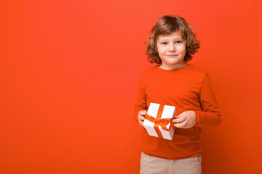 Shot of happy positive blond curly boy standing isolated over red background wall wearing red sweater holding gift box and looking at camera. Empty space