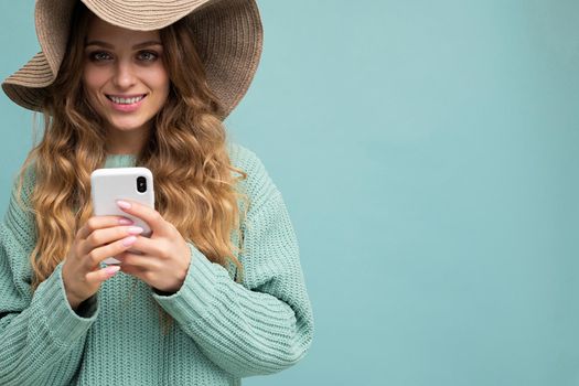 Closeup portrait photo of beautiful young blonde woman wearing blue sweater and hat standing isolated over blue background surfing on the internet via phone looking at camera. Copy space